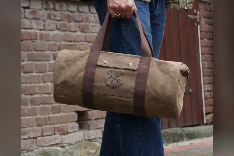 Duffle Bag aus gewachstem Canvas mit Siebdruck-Logo, in der Hand getragen vor einer Backsteinwand.