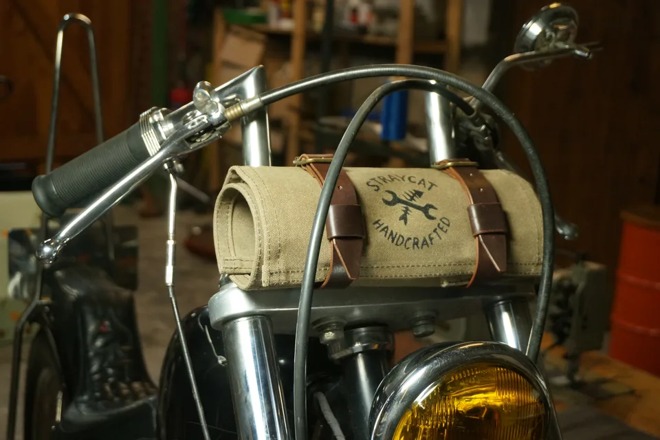 Tool Roll „Nine Lives“ in Desert Tan mit Lederriemen, festgeschnallt am Lenker eines Motorrads in der Werkstatt.