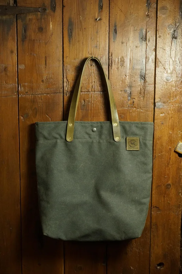 Tote Bag in Khaki mit Ledergriffen, hängend vor einer Holzwand.