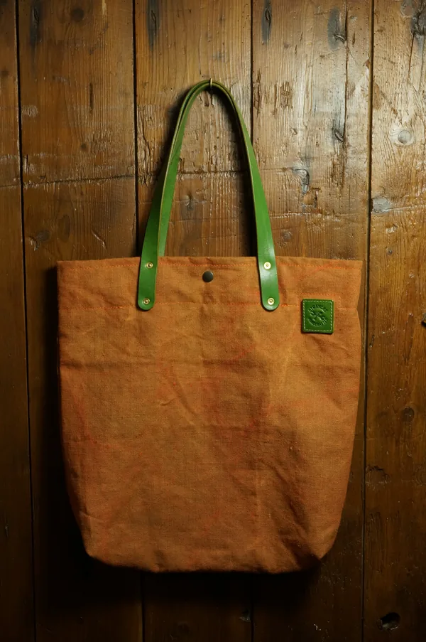 Tote Bag in Tent Orange mit grünen Ledergriffen vor einer Holzwand.